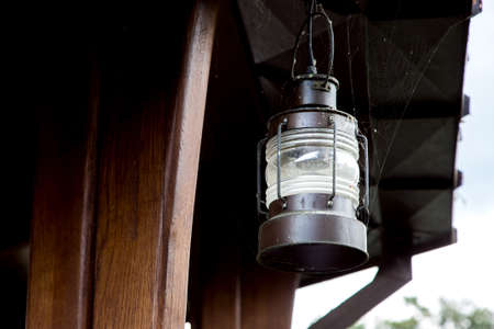 A retro pendant lamp of street lighting made of iron and a glass shade in the web is hanging on a wooden support of a brown gazebo.の写真素材