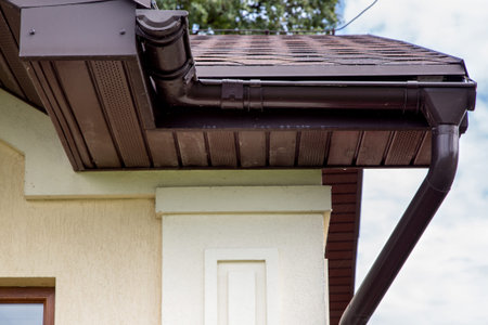 brown plastic drainage gutter on a roof with bituminous tiles close up of the house drainage pipes.
