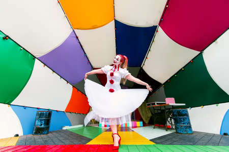 a girl in a white dress and a make-up clown on her face with red hair is standing on the street stage decorated with different bright colors, the image of a woman on the theme of Halloween.の写真素材