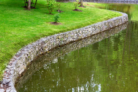 shore protection wall made of stone along the river, on the shore there is a lawn with green grass and bushes.の写真素材