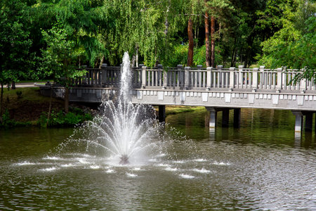 a fountain spraying water jets in a pond on a sunny day in a park with a stone bridge in the background.の写真素材