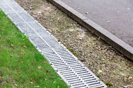 drainage system on the side of the asphalt road behind a gutter with an iron grid and green grass, close up.の写真素材