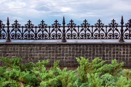 stone fence made of square tiles and with iron forged peaks with a pattern in a park with green bushes in the background a river and a cloudy sky.の写真素材