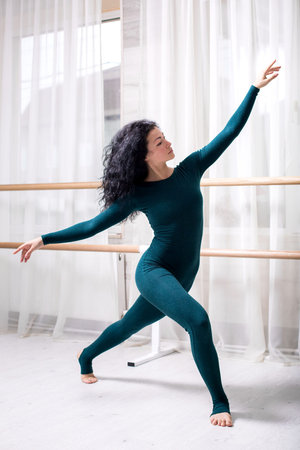 young brunette girl in green sportswear barefoot stands at the railing and poses in a bright gym.