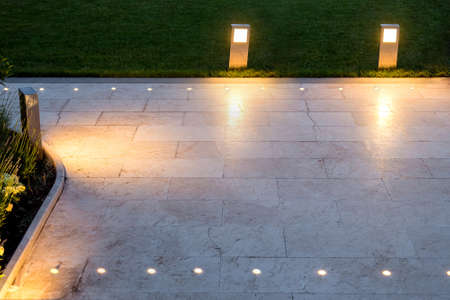 The pavement of marble tiles and a stone border area in the evening illuminated by ground lamp in a chrome shining a warm light, nobody.の写真素材
