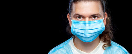 Male portrait a close-up of a face with a serious doctor expression in a blue medical mask isolated on a black background, a banner on a medical theme with copy space.の写真素材