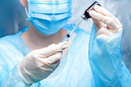 the doctor in mask hold in hands on gloves hold a glass bottle with a vaccine and a syringe with a...