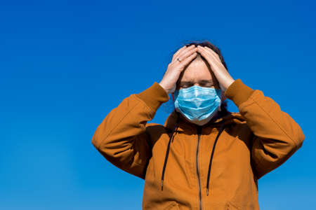 a man in a medical mask in an orange jacket holds his head in his hands from pain and stress due to a coronavirus pandemic, close-up with a copy space.の写真素材