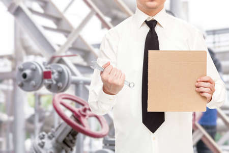 a worker in a shirt and tie holds a wrench, clenching his hand into a fist and a poster protesting against unemployment due to falling oil prices due to oil wars in the background of an oil refinery.の写真素材