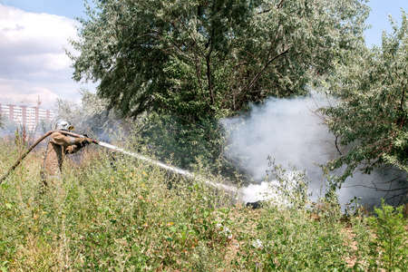 firefighter in a protective suit among the grass and trees extinguishes burning garbage in nature.の写真素材