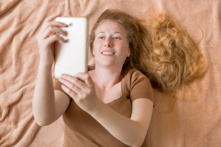 blonde woman with long hair lies on bed with a soft brown blanket and holds label tablet in hands,...
