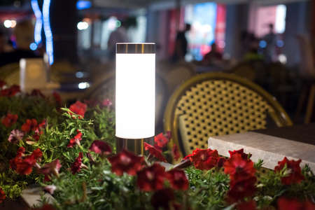 modern steel lantern with a white shade in a flowerbed with red flowers on the terrace of a street city restaurant night glow scene, nobody.の写真素材