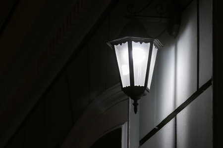 black iron retro lantern with transparent glass hanging on the wall of the facade of the building illuminating the architecture of the city at night, closeup glow detail side view, nobody.の写真素材