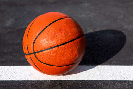 orange striped basketball ball stands on the white line marking the playing field with asphalt tarmac, sports object lit by sun light on playground, nobody.の写真素材