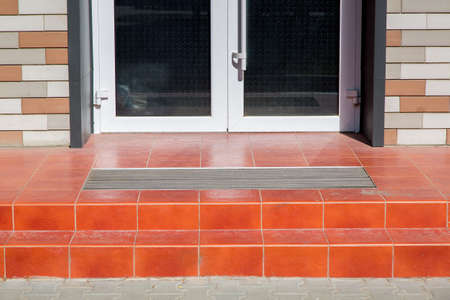 threshold with a paved red tile and a foot mat at the front door of a brick walled building on a sunny day, close-up step at the entrance door, nobody.の写真素材