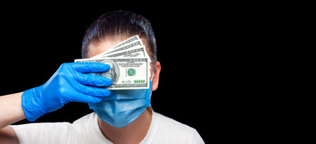 portrait of a doctor in a protective mask and sterile gloves holding a bribe covering his corrupt face close-up isolated on a black background.の写真素材