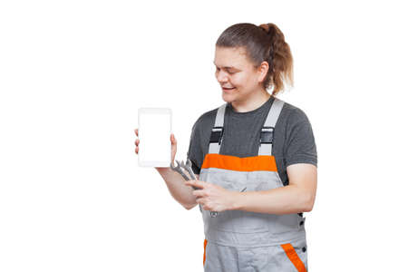 male mechanic in gray uniform with spanner indicates in his tablet with white screen mock up on theme of online service and remote renovation, man with tools isolated on white background.の写真素材