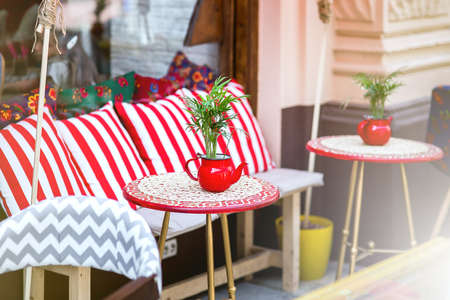 a red round table with a tablecloth and a flowerpot in the form of a teapot with a green plant by a chair with pillows, an outdoor cafe with empty seats, nobody.の写真素材