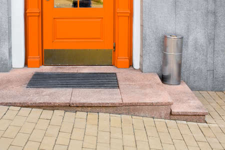 granite threshold with foot mat near orange wooden front door with iron trash can near building facade with gray cladding and yellow stone tile pavement close-up, nobody.の写真素材