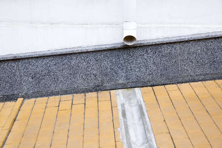 gray granite cladding on facade of a house wall with a drainage pipe and a concrete dish canal on a stone slab sidewalk to drain rainwater front view.の写真素材