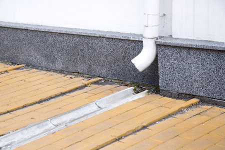 facade of a building wall with a white drainage pipe and a concrete dish channel on a stone slab sidewalk to drain rainwater, city architecture closeup drain system.の写真素材