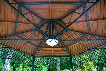 hanging lamp on the ceiling of the gazebo with an iron frame and wooden decorative inserts, a canopy in the backyard with an illuminator among plants close-up.の写真素材