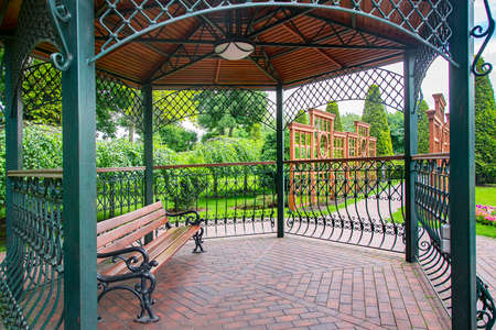 iron gazebo with ceiling lighting for relaxing on a wooden bench in the backyard with vertical pergola for growing plants in a park with bushes and trees on a green lawn parkland, nobody.の写真素材