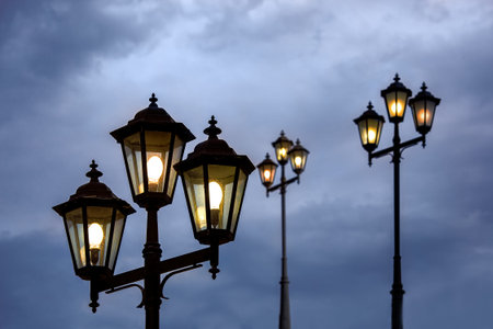 triple lantern head with transparent glass panels and electric bulb, iron street lighting glowing in the evening against a blue cloudy sky, retro style lamp fixture.の写真素材