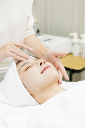 Relaxed Asian woman enjoying facial massage with closed eyes in beauty salon. Face Treatment. Beauty Injection Procedure for Skincare. Close up of Beautician Hands.の写真素材