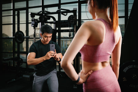 Young slim fitness Asian woman standing with personal trainer taking a photo by mobile phone in the gym. The athlete leads a healthy lifestyle. Cardio training for weight loss.の写真素材
