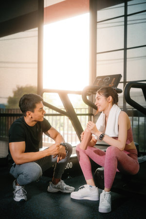 Young slim fitness Asian woman sitting with personal trainer conversation for a break in the gym. The athlete leads a healthy lifestyle. Cardio training for weight loss.の写真素材