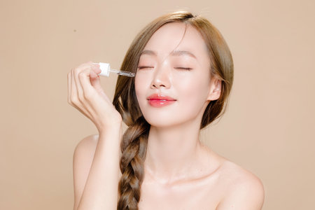 Young Asian woman long hair braid with natural makeup on face and clean fresh skin using dropper to apply serum on isolated beige background. Female model in studio. Facial treatment, Cosmetology.の写真素材