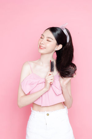 Cute Asian woman model gathered in ponytail with korean makeup style on face have plump lips and clean fresh skin wearing pink camisole combing hair on isolated pink background.の写真素材