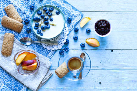 Healthy Breakfast with blueberries and banana yogurt, biscuits, marmalade, coffee and peaches over a light wooden background.の写真素材