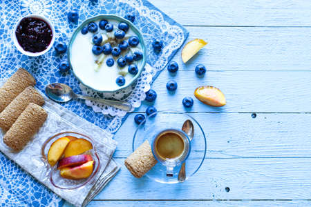 Healthy Breakfast with blueberries and banana yogurt, biscuits, marmalade, coffee and peaches over a light wooden background.の写真素材