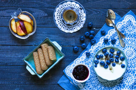 Healthy Breakfast with blueberries and banana yogurt, biscuits, marmalade, coffee and peaches over a light wooden background.の写真素材