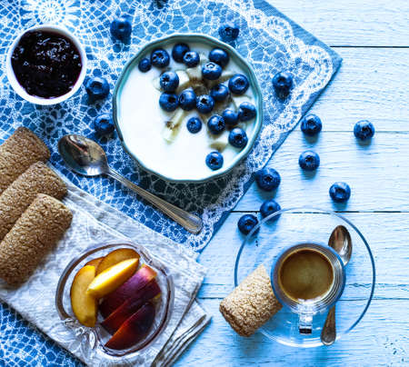 Healthy Breakfast with blueberries and banana yogurt, biscuits, marmalade, coffee and peaches over a light wooden background.の写真素材
