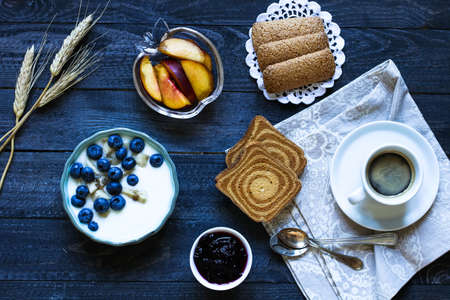 Healthy Breakfast with blueberries and banana yogurt, biscuits, marmalade, coffee and peaches over a light wooden background.の写真素材