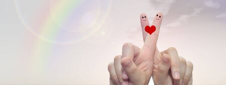 Conceptual fingers in love, with a painted heart shape over rainbow sky background bannerの写真素材