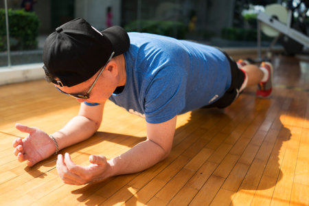 Man wearing glasses and a cap. He open his hands while doing plank exercise in a gym with wooden...