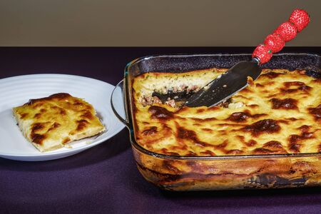 Greek moussaka dish recipe preparation. Ready meal with served portion next to transparent bowl of baked mousaka with bechamel white sauce & fried chopped potatoes above ground chopped beef meat.の写真素材