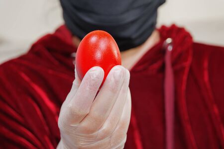Person holding red Easter hard-boiled egg before camera. Unidentified female wearing black face mask and white protective gloves displays painted egg.の写真素材