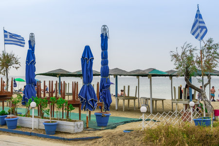 Neoi Epivates, Greece Empty Beach bars prepare to open for summer season. Closed outdoor bar restaurants with Greek flag, stacked tables, bamboo sun umbrellas & bathers on sand by sea.のeditorial素材