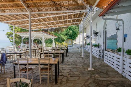 Chalkidiki, Greece - July 30 2020: Almost empty tavern chairs & tables after new covid-19 measures. Restaurant outdoor seating area by the sea with a few customers in Kryopigi, Kassandra peninsula.のeditorial素材