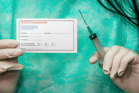 Chemist with protective green suit holds a covid-19 vaccination card. Medical concept of female with...