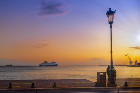 Thessaloniki, Greece - March 6 2021: Blue Star Mykonos passenger ship sailing to city seaport at sunset. Evening view of commercial boat entering calm sea harbor, with Hellenic flag waiving on a craneのeditorial素材