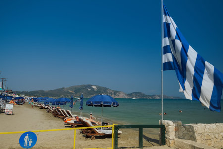 Zakynthos, Greece - July 9 2021: Mediterranean terrain organized beach with Greek flag and bathers....