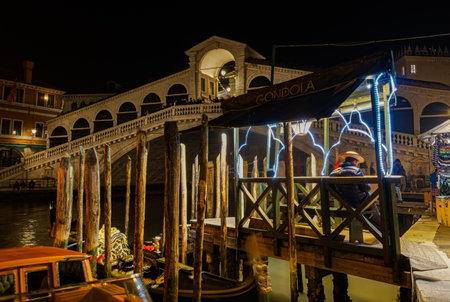 Venice, Italy - February 22 2023: night view of Ponte di Rialto, Rialto pedestrian Bridge with a gondolier in the front ground, next to wooden wharf pilings.のeditorial素材