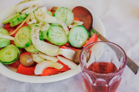 A small glass of red wine next to a dish of served salad with sliced cucumber, tomatoes, onions and olives.の写真素材