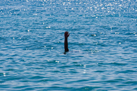 Drowning man concept, with a male raised hand with an open palm visible waving for help above a blue sea with low waves.の写真素材